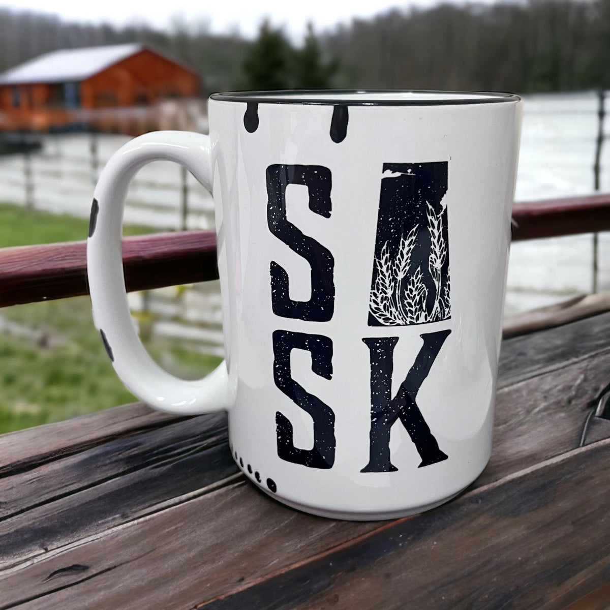 White ceramic farmhouse mug with large distressed SASK lettering and wheat stalk design, on a wooden deck with a barn